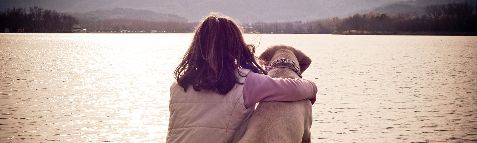 A girl hugging a dog as they look over a lake