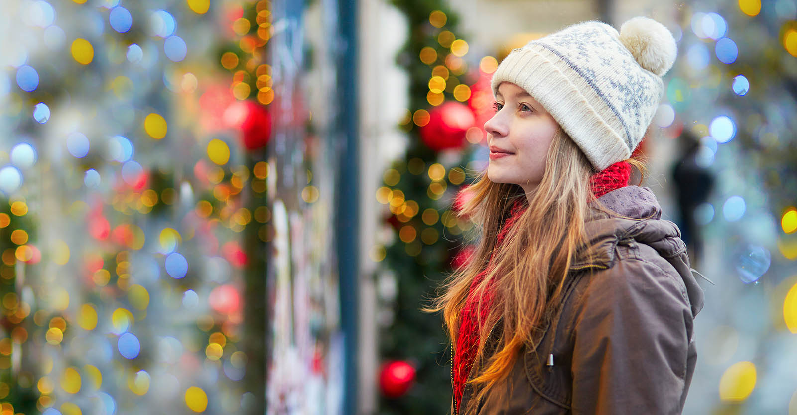 A woman in her winter coat and hat looking into a window with Christmas ornaments.